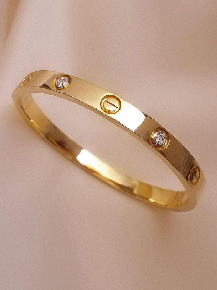 Yoniq.woman gold-tone metal bangle bracelet with crystal inlays and engraved screw-head motifs on a silk background.