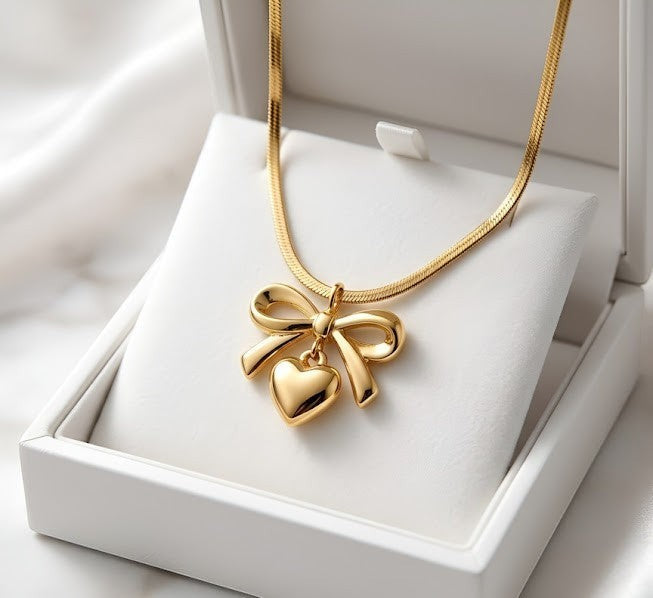 Yoniq.woman gold-tone necklace featuring a polished heart and bow pendant on a sleek herringbone chain in a gift box.