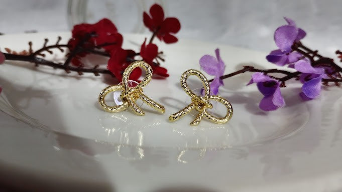 Yoniq.woman gold-tone bow stud earrings with a twisted rope texture on a white plate with red and purple flowers.