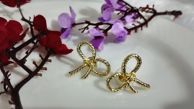 Yoniq.woman gold-tone bow stud earrings with a twisted rope texture on a white plate with decorative flowers.