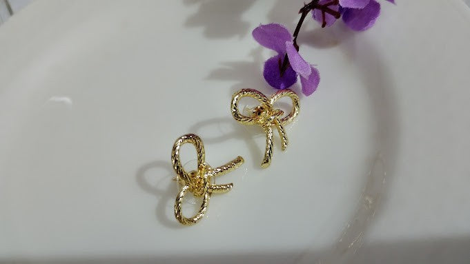 Yoniq.woman gold-tone bow stud earrings with a twisted rope texture on a white plate with purple floral accents.