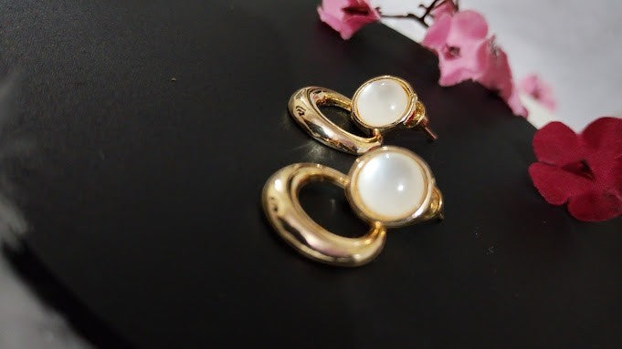 Yoniq.woman gold-tone circular drop earrings with smooth white cabochon studs on a black surface with pink flowers.