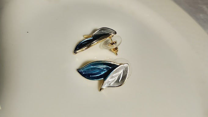Yoniq.woman leaf-shaped stud earrings with blue and white enamel and gold-toned metal edges on a white surface.