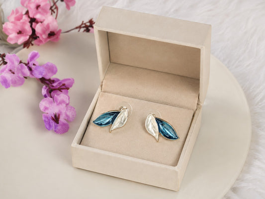Yoniq.woman leaf-shaped stud earrings with blue and white enamel and gold-toned edges in a beige jewelry box.
