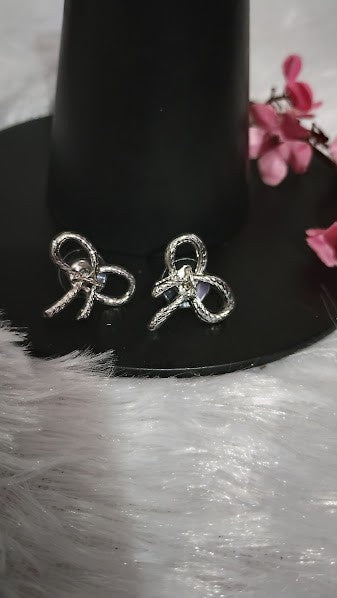 Yoniq.woman silver-tone textured bow stud earrings with a delicate knotted ribbon design on a black display stand.