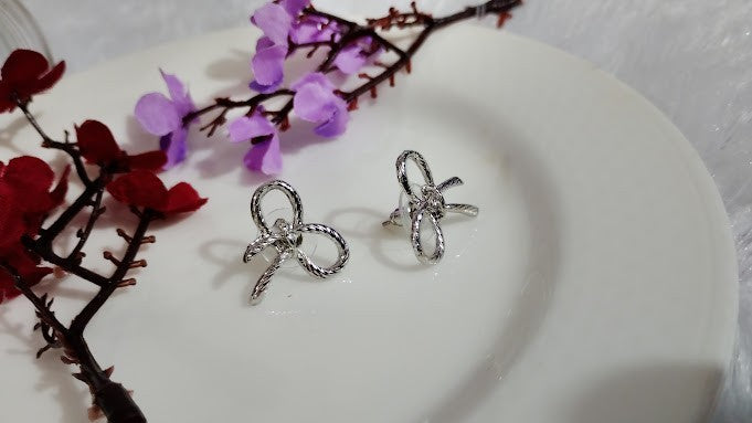 Yoniq.woman silver-tone bow stud earrings with a textured metallic ribbon design resting on a white plate with flowers
