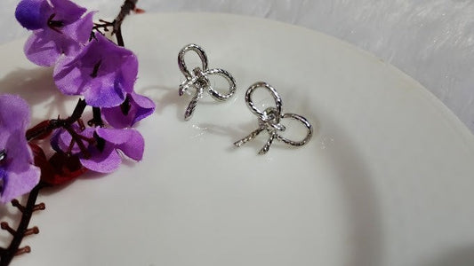 Yoniq.woman silver-tone textured bow stud earrings with a delicate knotted ribbon design on a white plate with flowers