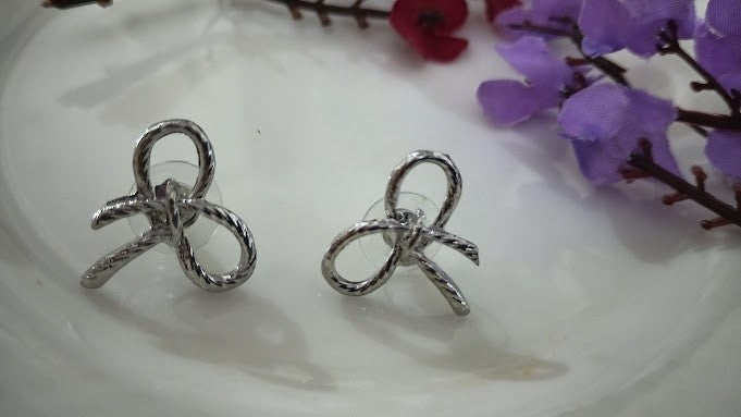 Yoniq.woman silver-tone bow stud earrings with a textured metallic ribbon design on a white plate with purple flowers.